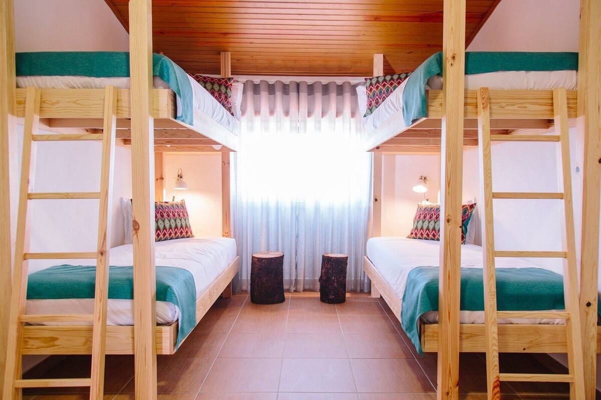 Une chambre avec deux lits superposés, chacun recouvert d’une couverture verte et agrémenté d’oreillers à motifs. On y trouve deux petits tabourets en bois et une fenêtre ornée de rideaux légers.