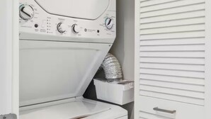 A compact, white laundry room with a front-loading washer and dryer, a small shelf with a rolled-up towel, and a door with horizontal louvered panels. - Union Station Stay | Spacious Unit w/ Kitchen | Walk to Parks & Museums (Pittsburgh)