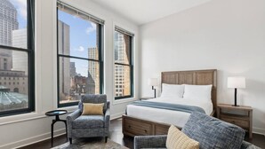 Ett hotellrum med en stor säng, två fåtöljer, ett litet bord och utsikt över stadens byggnader genom stora fönster.