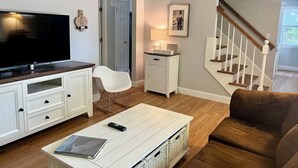 A living room with a flat-screen TV, white furniture, a staircase, and a door leading to another room. - Marvelous 4-bedroom, 2-bath home with AC and private backyard close to the beach (Narragansett)