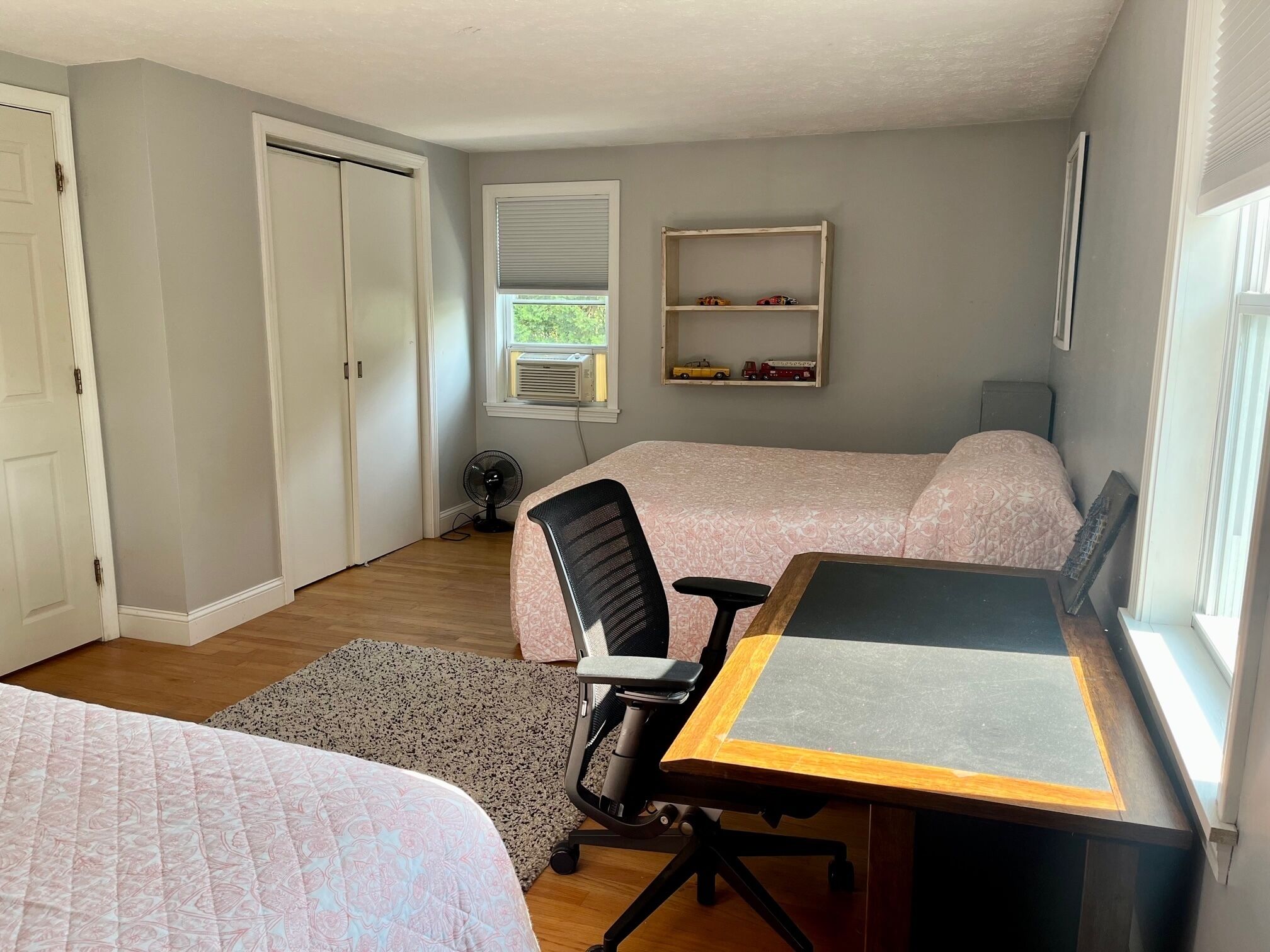 Een slaapkamer met twee bedden, een bureau, een stoel, een ventilator en een plank met speelgoed.