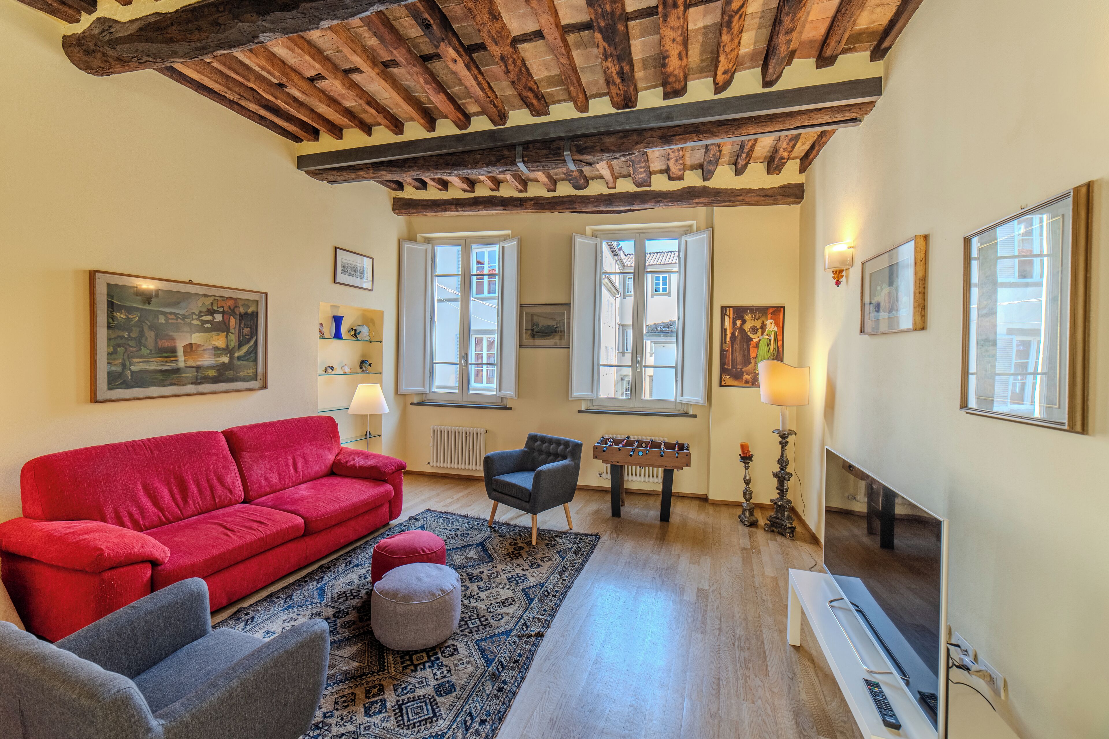 A cozy living room with a red sofa, grey armchairs, a wooden coffee table, and a foosball table. There are two windows with white shutters, a fireplace, and a television.