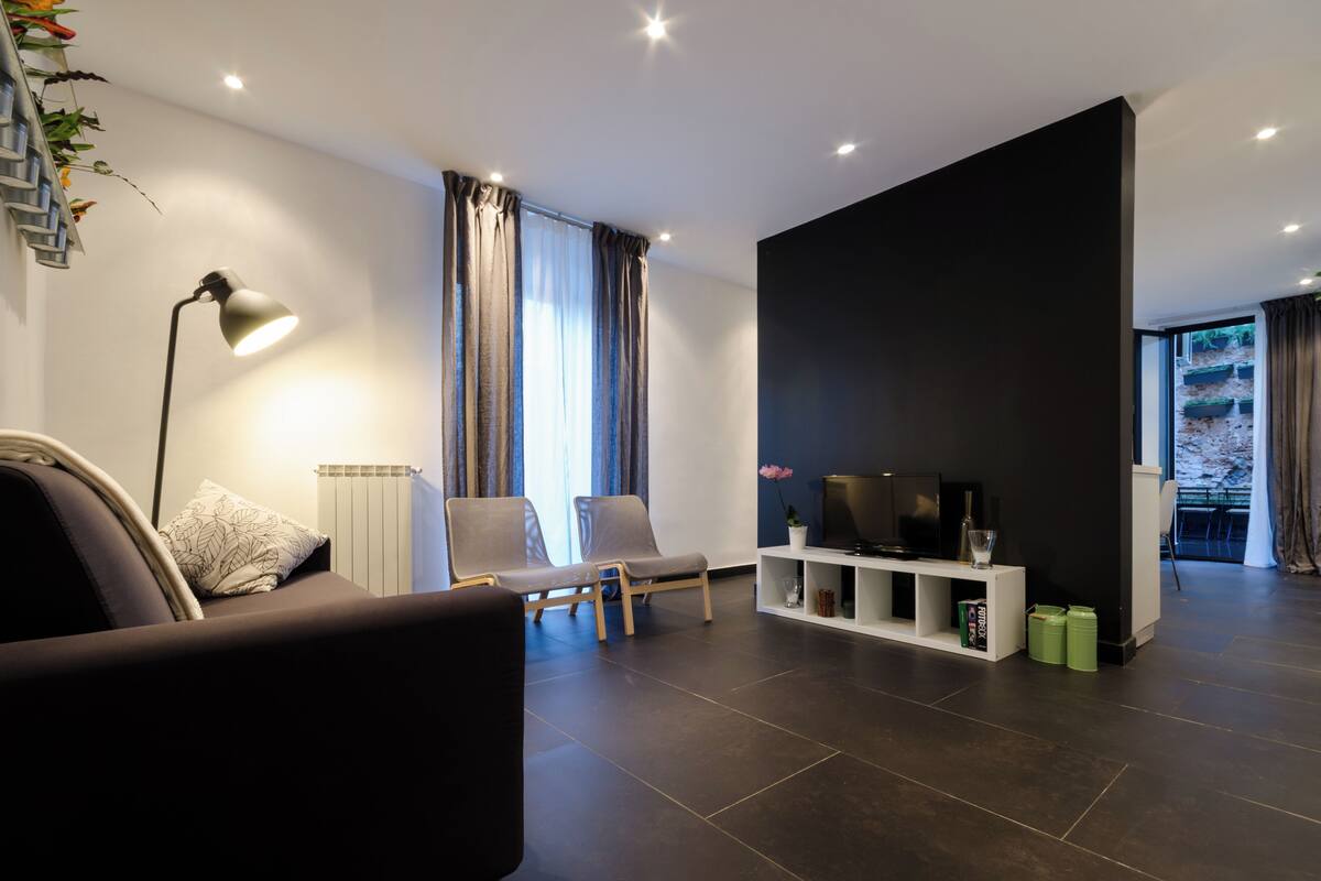 A modern living room with a sofa, two armchairs, a TV on a white stand, and a large black accent wall.