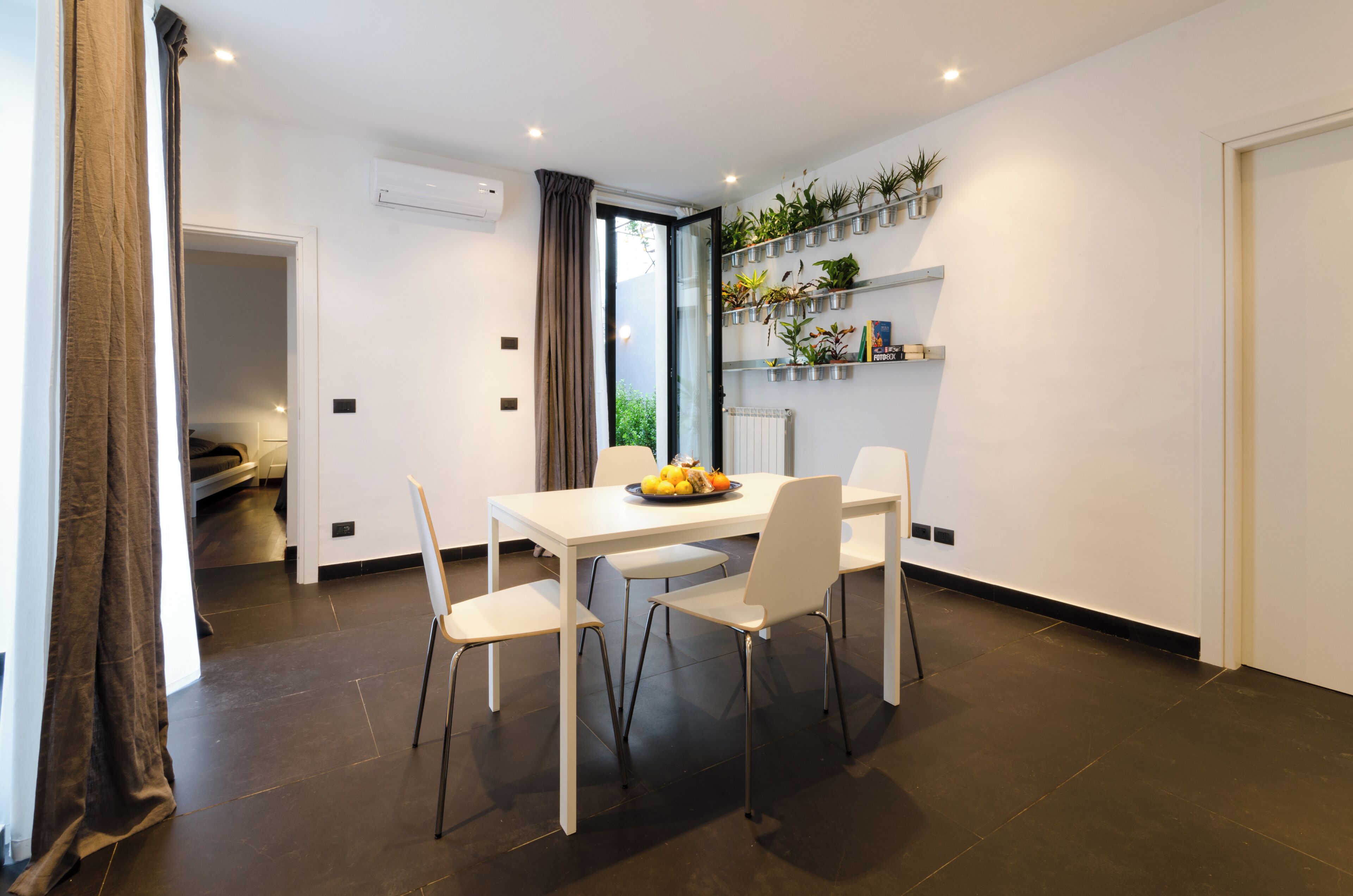 A modern dining area with a white table, four chairs, and a fruit bowl. There are potted plants on shelves and a view of greenery outside.