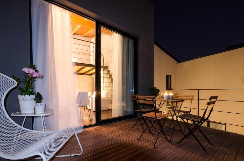 Un balcón exterior con terraza de madera, una silla blanca, una mesita y una parrilla.