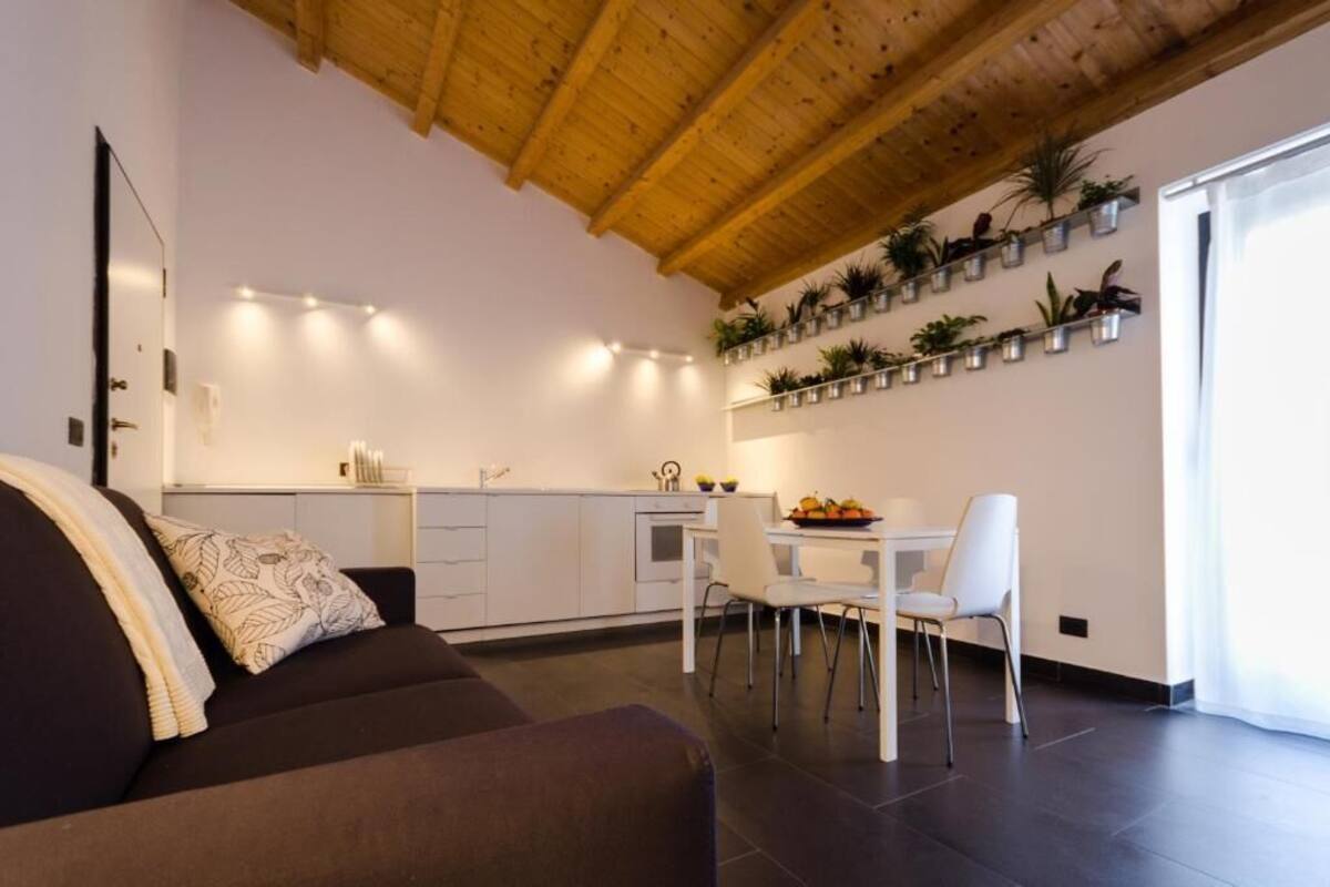 A modern living room with a dark brown sofa, a white dining table with chairs, and a kitchen area with white cabinets and a wooden ceiling.