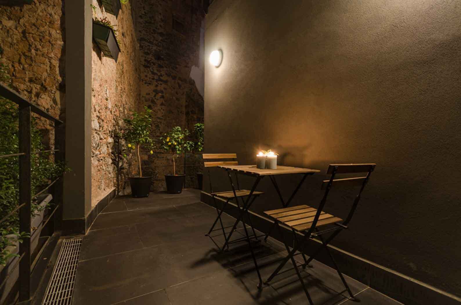 Une terrasse extérieure avec une table et des chaises, des plantes en pot et un luminaire fixé au mur.