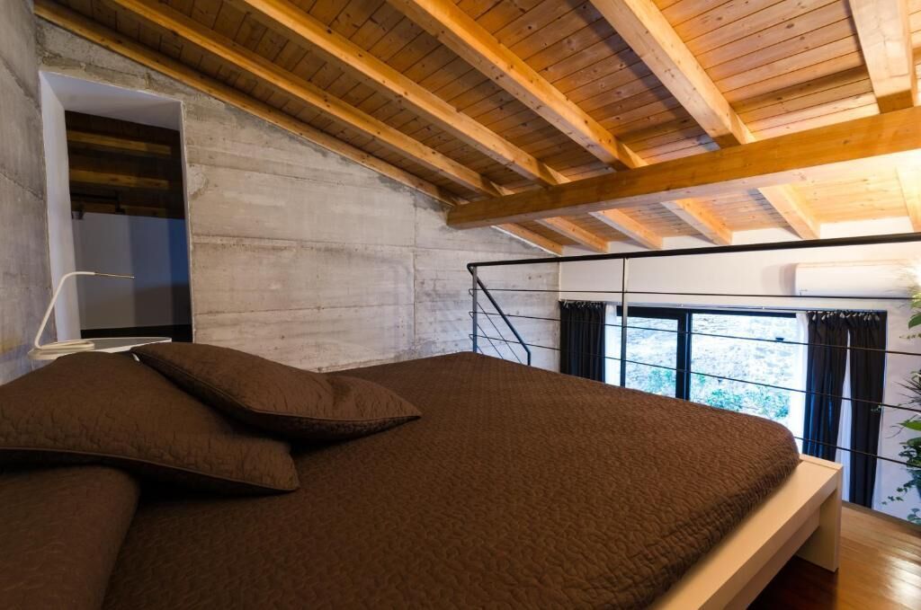 Un dormitorio con techo de madera, cama con ropa de cama marrón, ventana con cortinas y vista a zonas verdes.