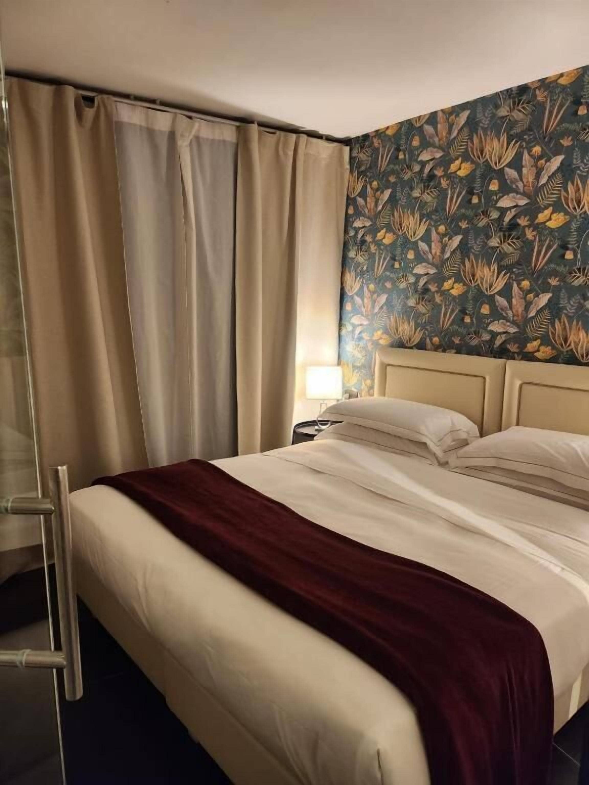 Habitación de hotel con cama, cortinas, mesita de noche y un papel tapiz floral.