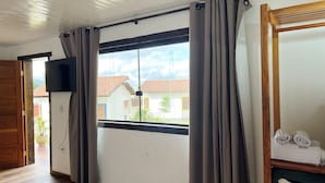 A room with a large window, wooden flooring, a television mounted on the wall, and a wooden shelf with towels. - Pousada Villa Salgueiro (Ouro Preto)
