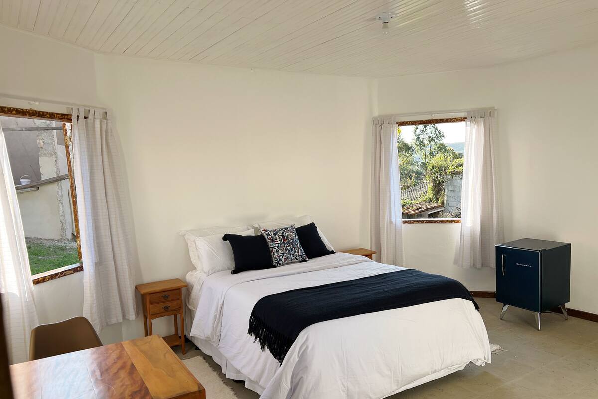 A bedroom with a bed, a wooden table, a small cabinet, and two windows with curtains.