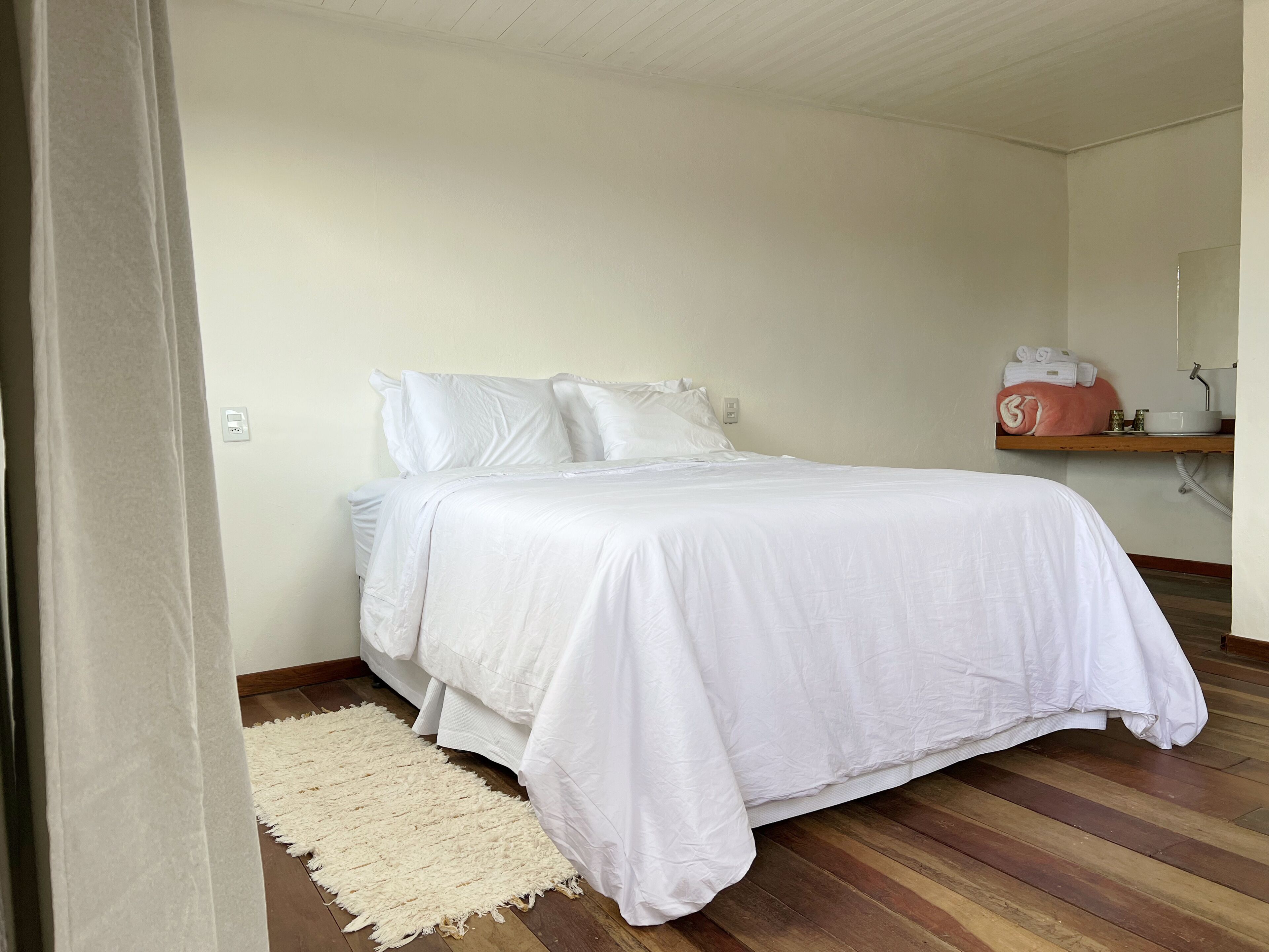 A bedroom with a bed, a white rug, a wooden floor, a shelf with towels, and a mirror.