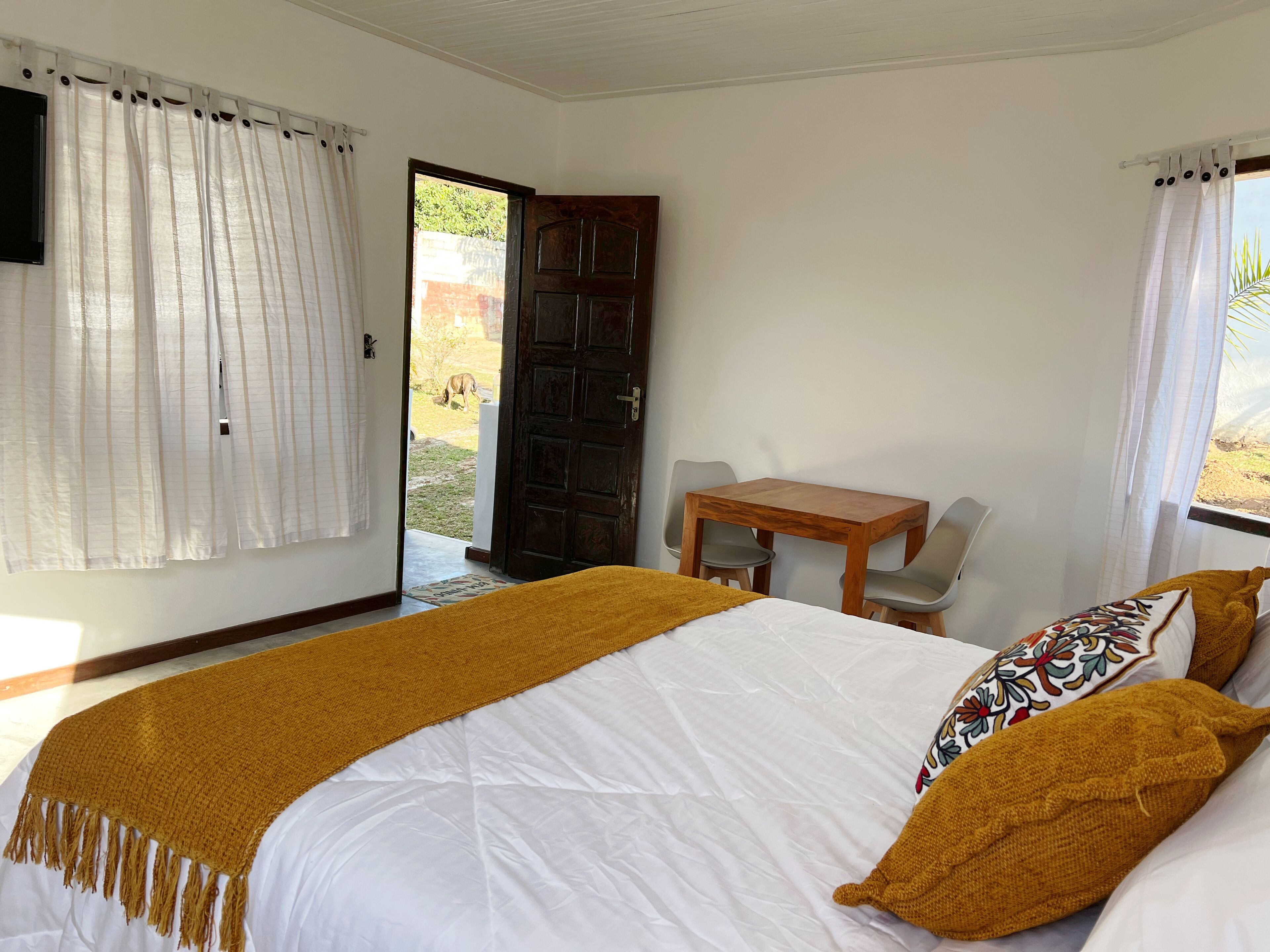 Una habitación de hotel con una cama, una mesa de madera con dos sillas, una puerta y una ventana con cortinas.