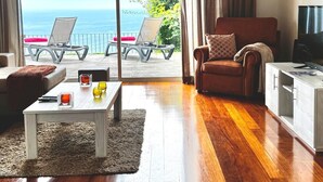 Un salon moderne avec un plancher en bois, un canapé, une table basse et un meuble pour la télévision. Il y a un balcon avec vue sur la mer.