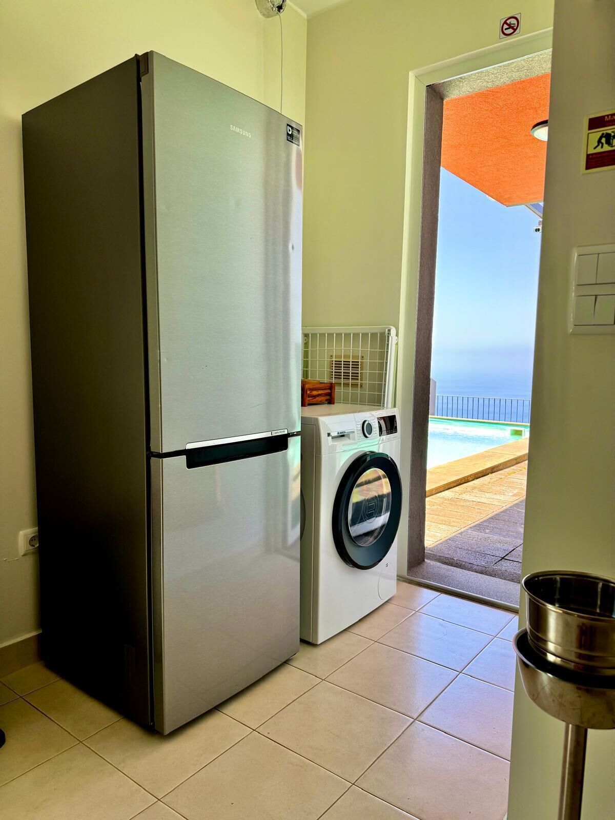 A compact kitchen with a stainless steel refrigerator, a washing machine, and a view of a pool through an open door.