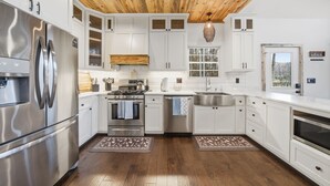A modern kitchen with white cabinets, stainless steel appliances, a wooden ceiling, and a window overlooking trees. - Creeks n' Peaks - Ellijay, GA (Ellijay)