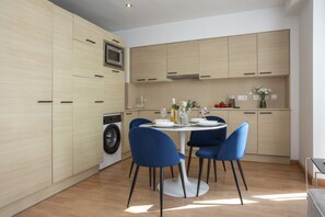 A modern kitchen with light wood cabinets, a built-in washing machine, a dining table with blue chairs, and a small countertop with a vase of flowers. - La Pergola 2B - Double Studio Exterior (Castelló de la Plana)