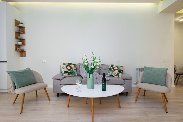 Un salon moderne avec une table basse ronde blanche, deux fauteuils, un canapé agrémenté de coussins à motifs, et un vase avec des fleurs et une bouteille.