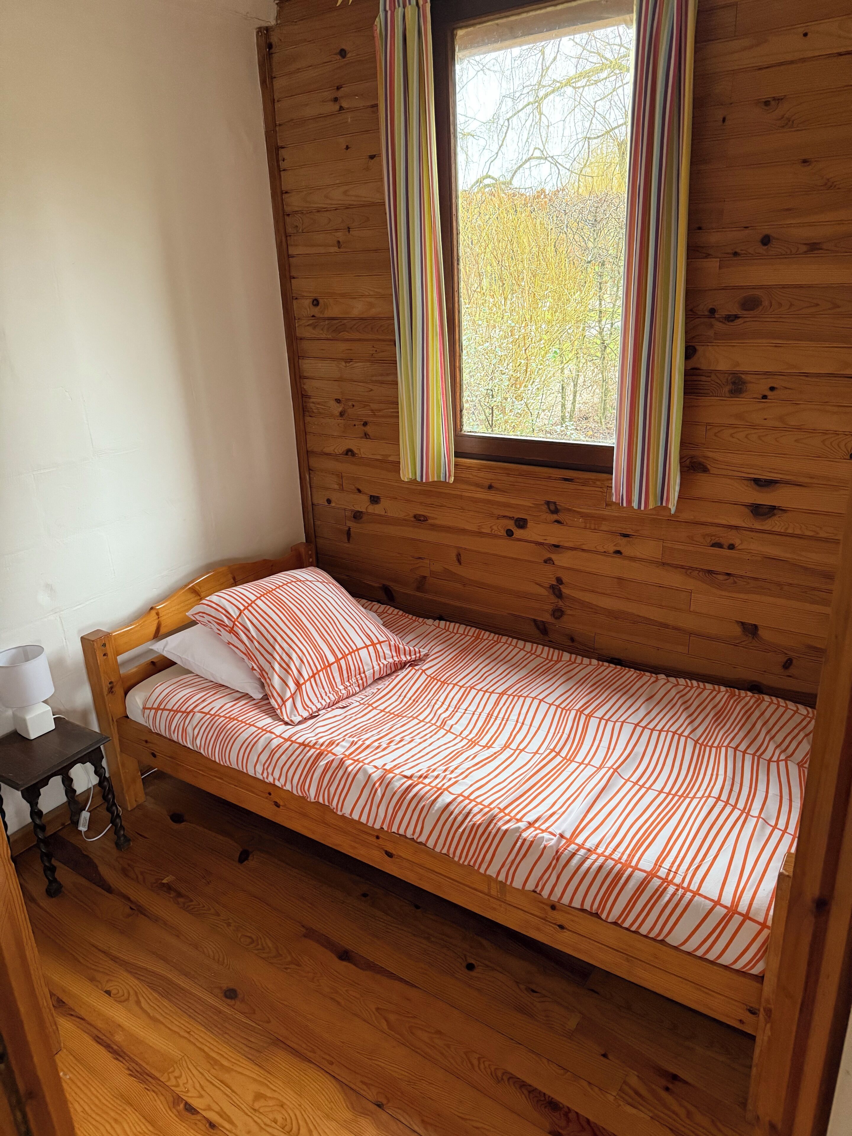A small wooden room with a single bed, a window with curtains, and a nightstand with a lamp.
