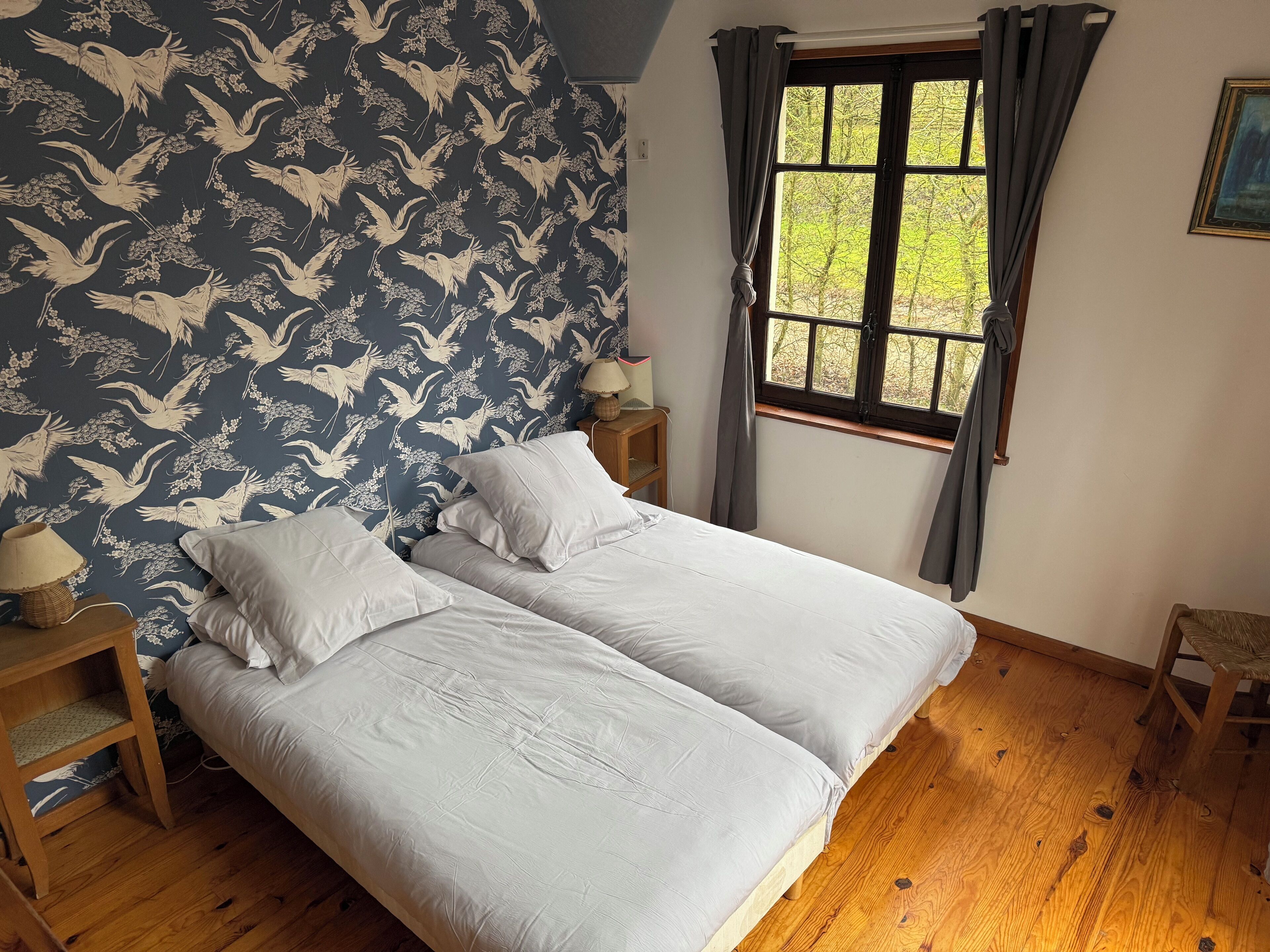 Una habitación con una cama, una mesita de noche, un ventanal con cortinas y un papel tapiz con diseño de pájaros.