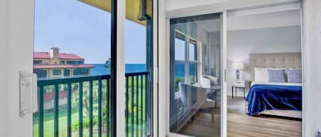 A room with a large sliding glass door leading to a balcony with a view of the sea and a building across the water.