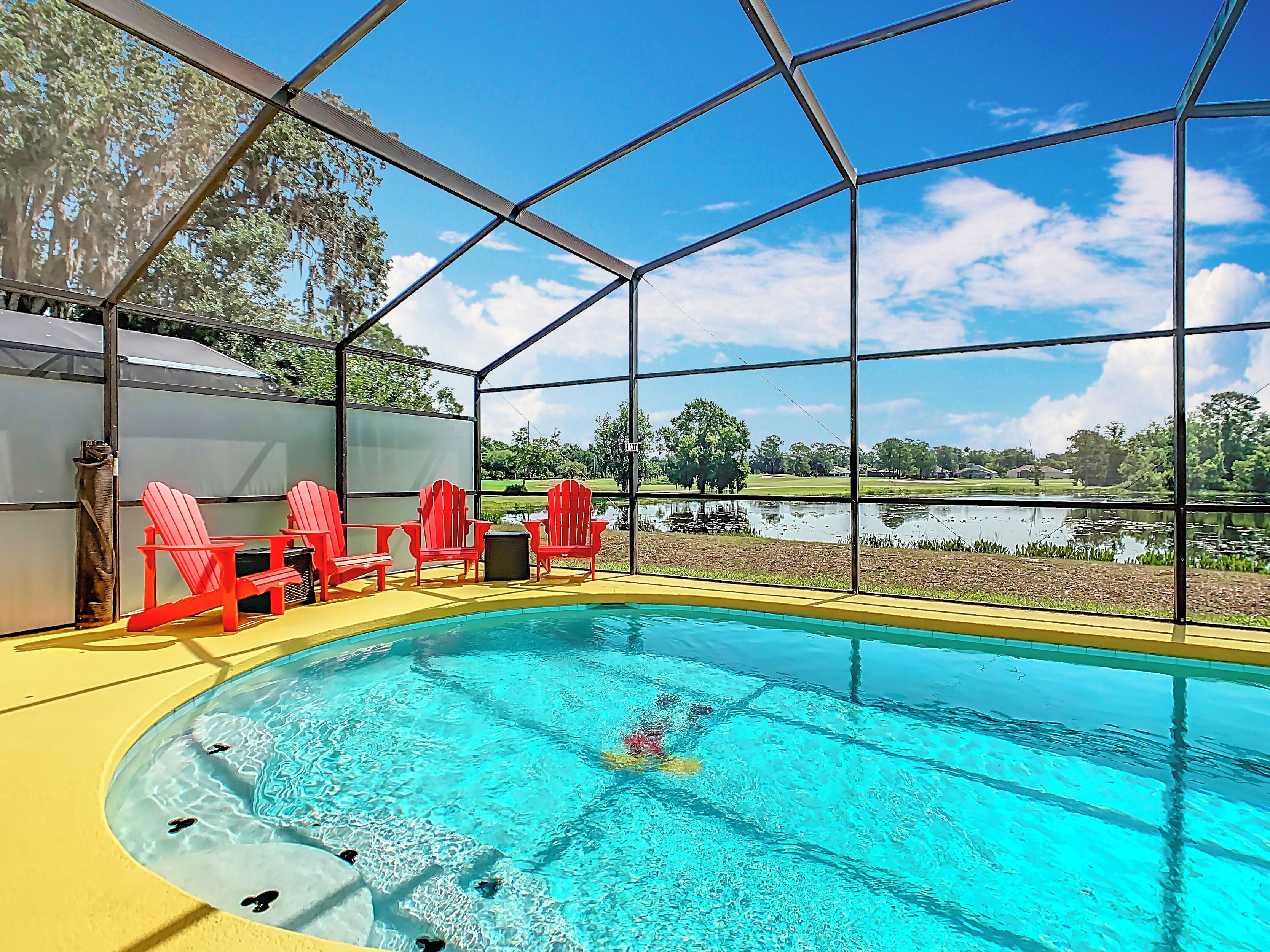 A pool area with a clear view of a lake and trees, surrounded by red chairs and a covered seating area.