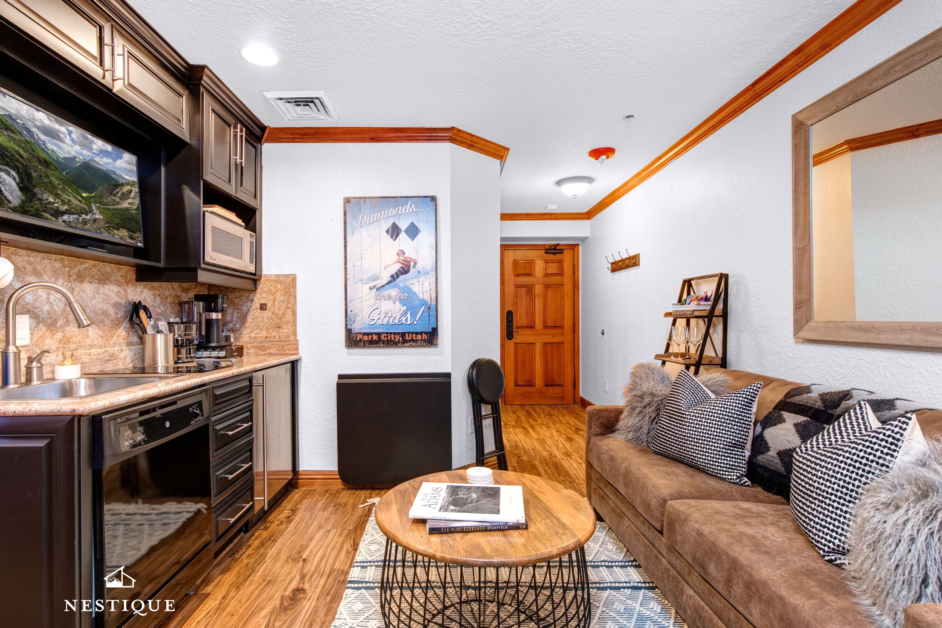 A modern kitchen with dark cabinetry, a built-in microwave, and a large window. There is a dining area with a brown sofa and patterned cushions. A poster on the wall advertises a park in Utah.