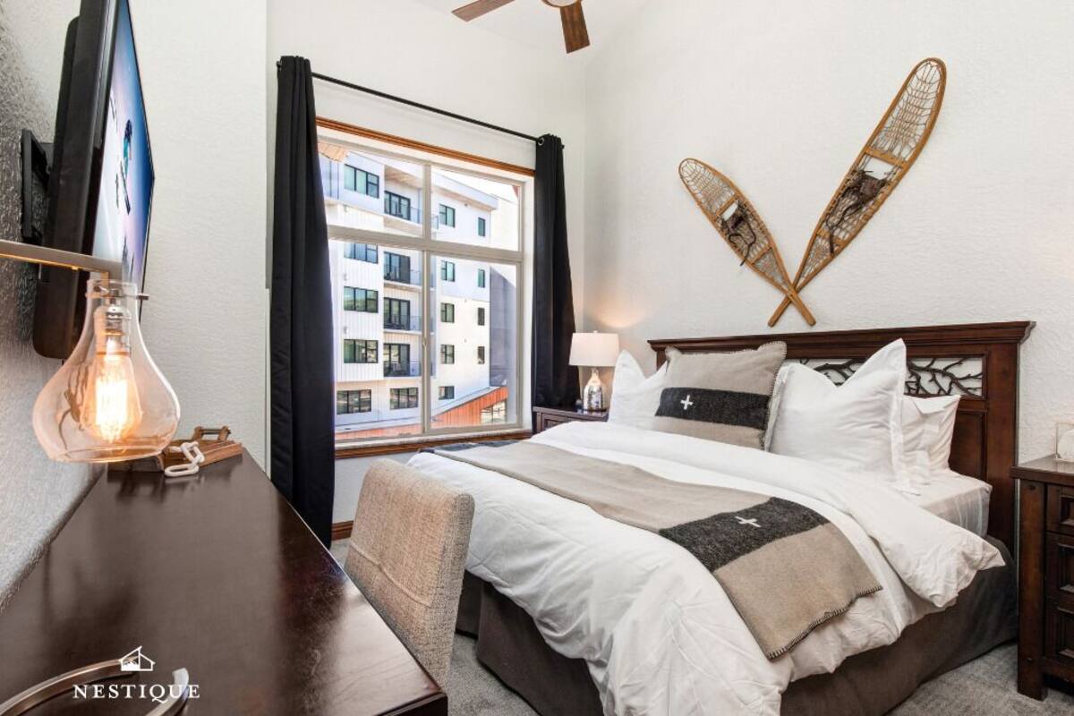 A hotel room with a bed, a desk with a lamp, a large window with curtains, and a wall-mounted decorative item.