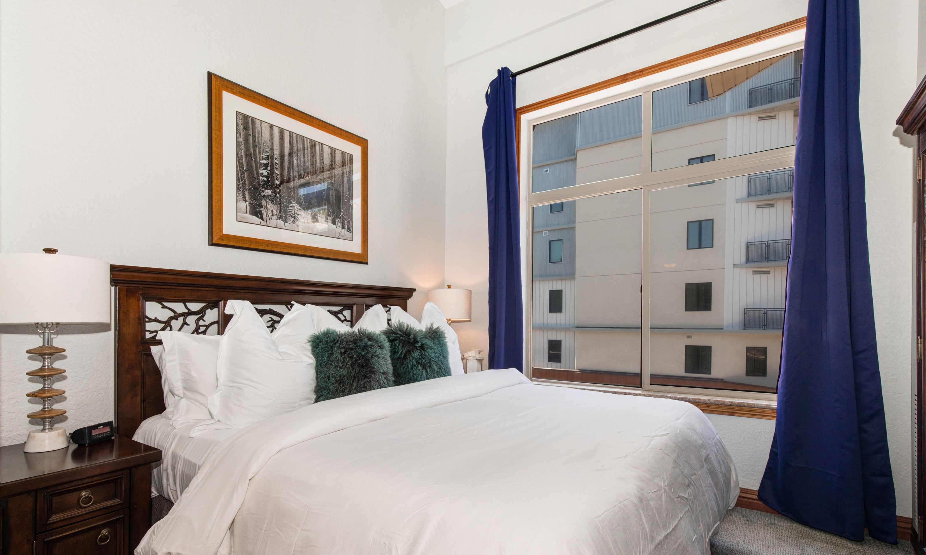 Un lit bien fait, avec du linge de lit blanc et des coussins verts, un tableau encadré au mur et une fenêtre avec des rideaux bleus.