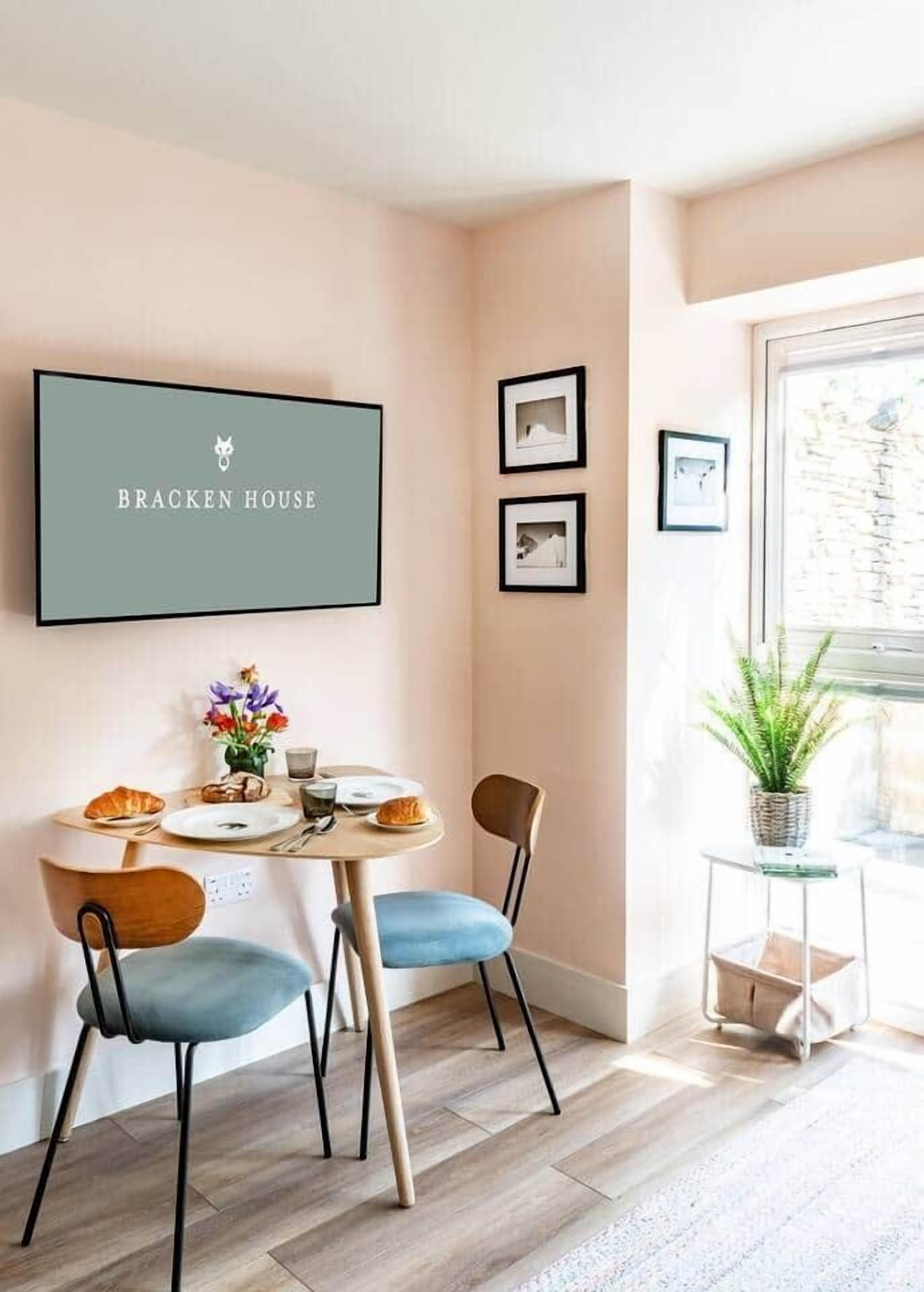 A dining area with a table set for two, chairs, a framed sign above the table, and a small potted plant on a side table.