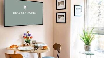 A dining area with a table set for two, chairs, a framed sign above the table, and a small potted plant on a side table.