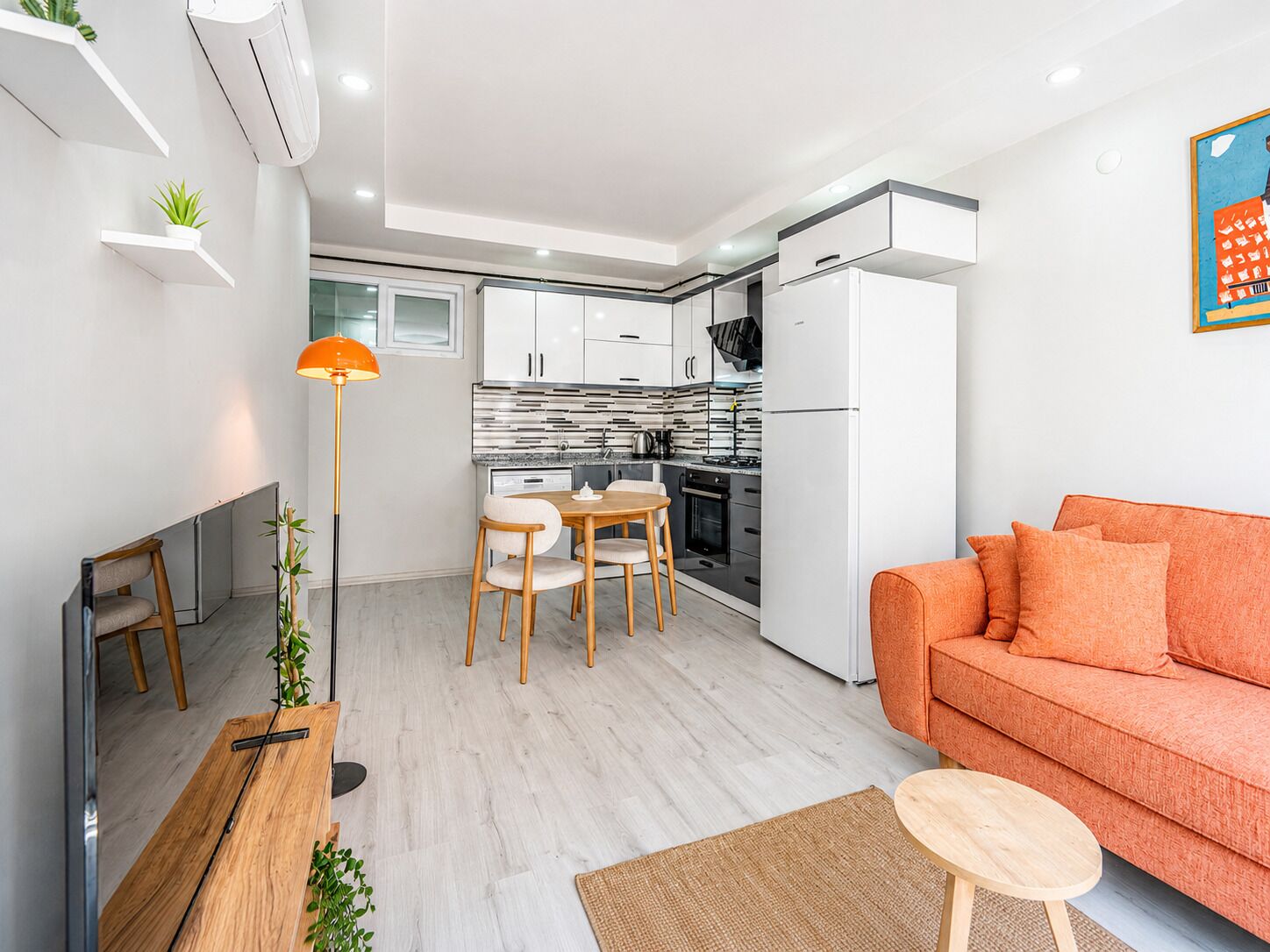 Una cocina moderna con armarios blancos, una zona de comedor con mesa y sillas de madera, un sofá y una lámpara de pie.