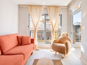 En moderne stue med sofa, stol, stort vindue med gardiner og udsigt til en balkon.