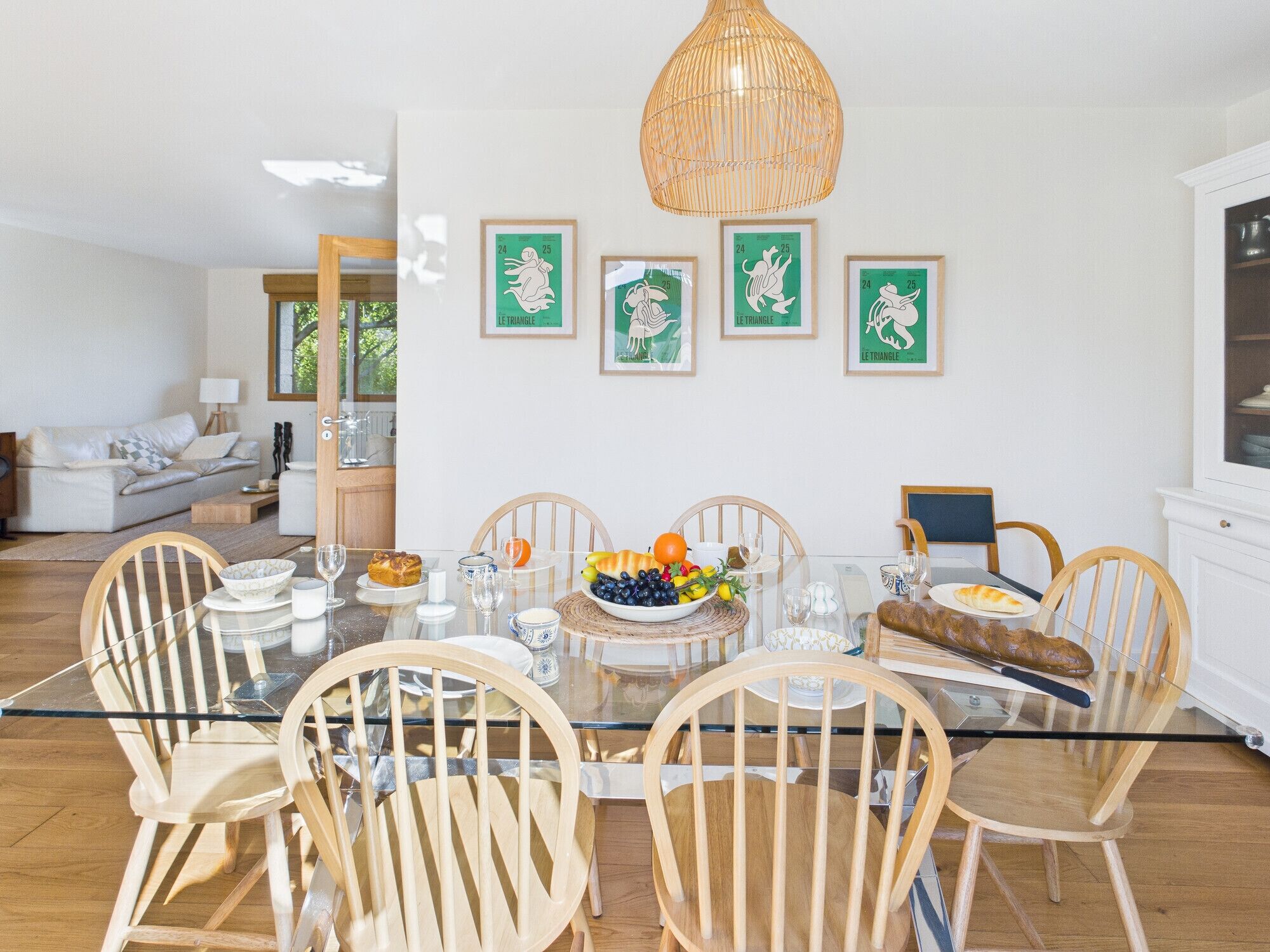 Uma sala de jantar com mesa de vidro, cadeiras de madeira e luminária pendente de vime.