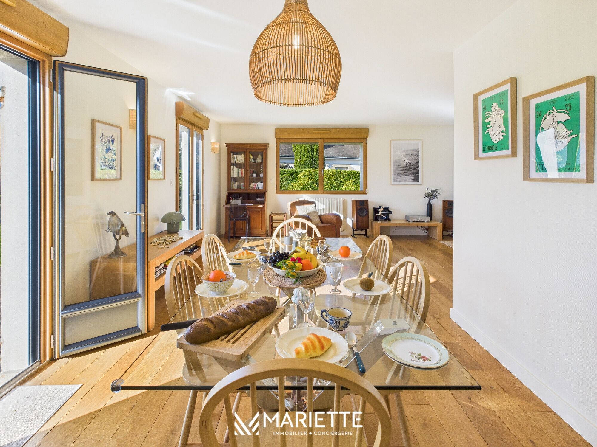 Un comedor con mesa de vidrio, sillas de madera y diversos alimentos. Se observa una estantería, una ventana con vistas a la vegetación y una lámpara colgante.