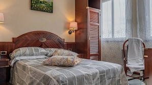 Ein ordentlich bezogenes Bett mit gemusterten Bettwaren, ein hölzernes Kopfteil, ein Nachttisch, eine Wandlampe, ein Kleiderschrank und ein Stuhl mit Handtuch.