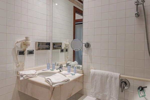 Une salle de bain avec un mur recouvert de carreaux blancs, un lavabo avec un miroir, un porte-serviettes et un espace douche.