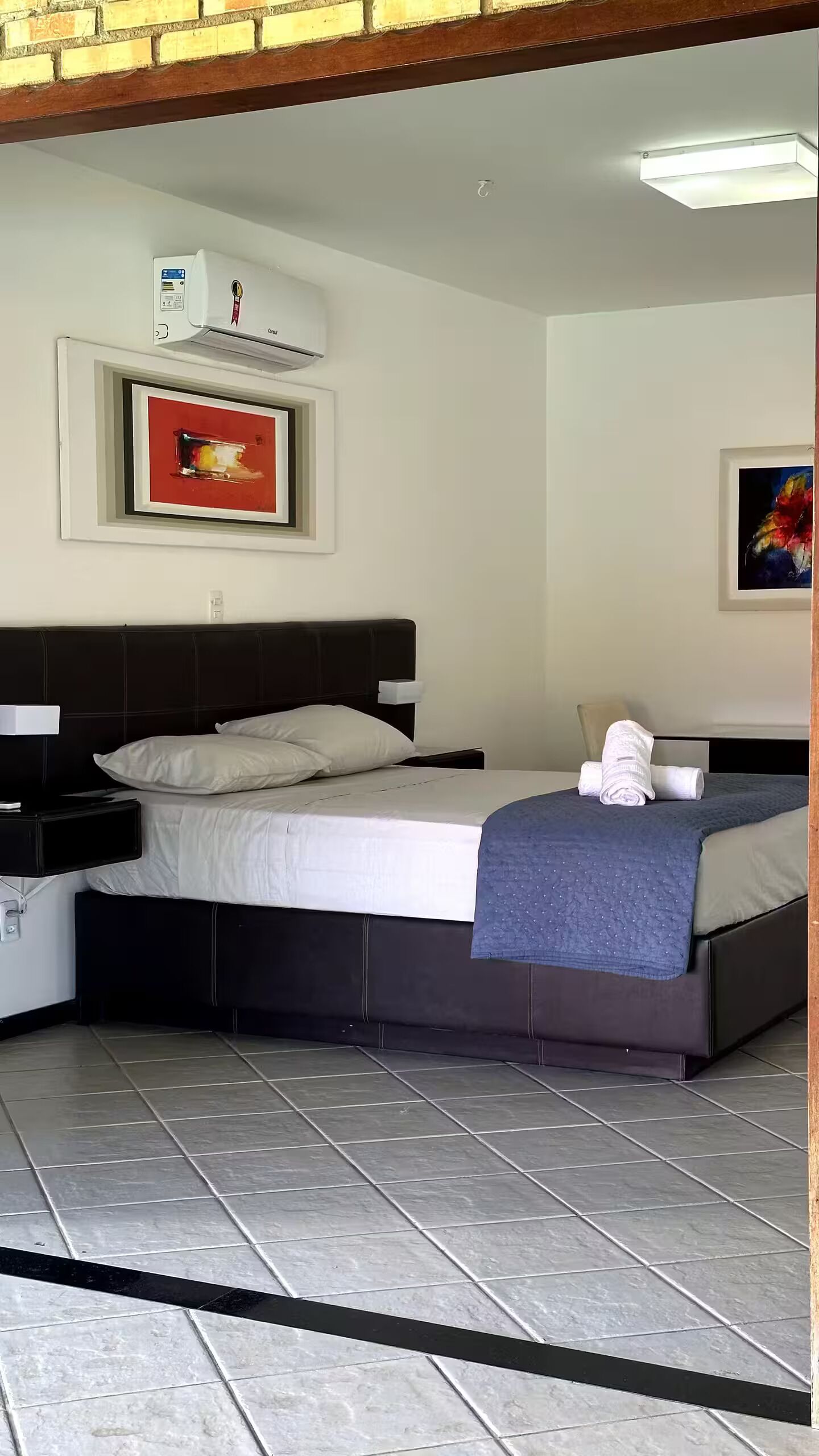 A neatly arranged hotel room with a bed, bedside tables, a nightstand, a wall-mounted air conditioner, and two framed artworks.