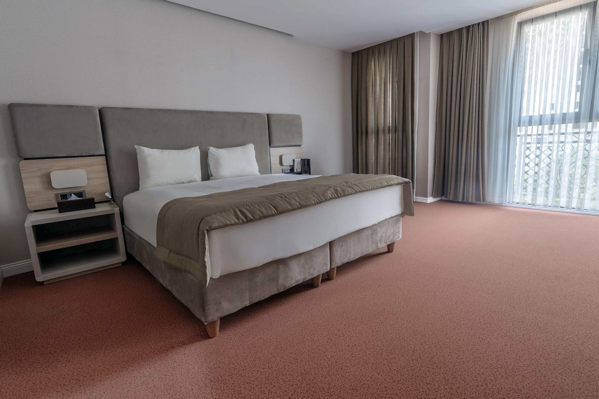 A modern hotel room with a large bed, a bedside table, and a view of the outdoors through sheer curtains.