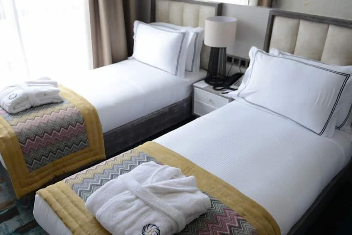 A hotel room with two beds, each with a pillow and a blanket. There is a bedside table with a lamp and a phone.