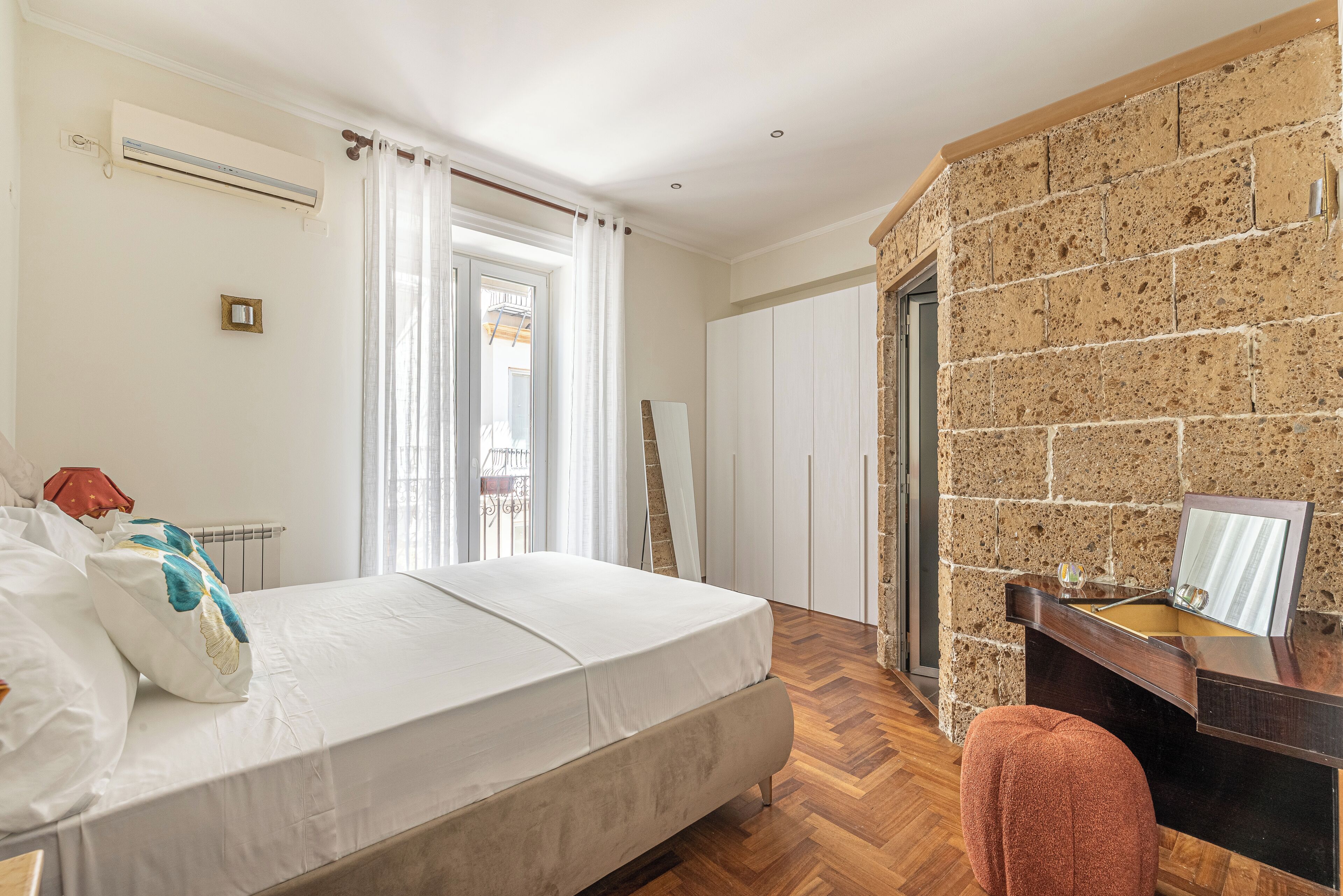 A bedroom with a bed, a wooden desk, a mirror, and a stone wall.