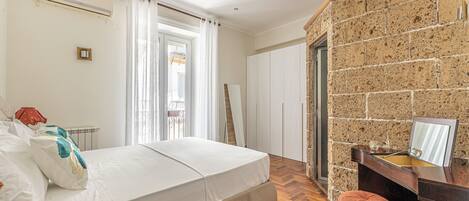 A bedroom with a bed, a wooden desk, a mirror, and a stone wall.