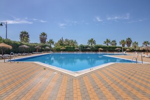 Une piscine entourée de palmiers, avec des chaises longues et des parasols, sous un ciel dégagé.