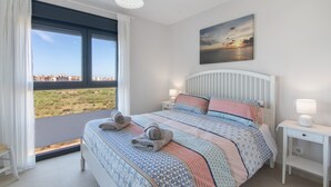 A bedroom with a large window, a bed with patterned bedding, a bedside table with a lamp, and a painting of a sunset. - Apartment 'Los Flamencos' with Sea View, Wi-Fi and Air Conditioning (Isla Canela)