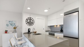 A modern kitchen with white cabinetry, a stainless steel refrigerator, a large island with seating, and a wall-mounted clock. - Apartment 'Los Flamencos' with Sea View, Wi-Fi and Air Conditioning (Isla Canela)