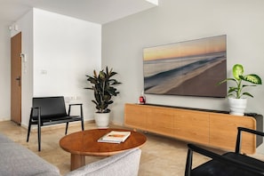 A modern living room with a round wooden coffee table, a black sofa, a black armchair, a wooden cabinet, and a large wall-mounted TV.