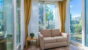 A modern room with a beige sofa, a small wooden table with a plant, and large windows with yellow curtains. -  Hotiday Lodge Cecina (Cecina)