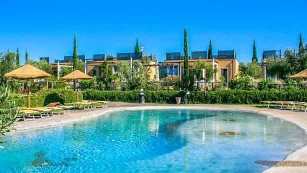 A swimming pool with lounge chairs and umbrellas, surrounded by well-maintained hedges and trees, with modern buildings in the background. -  Hotiday Lodge Cecina (Cecina)