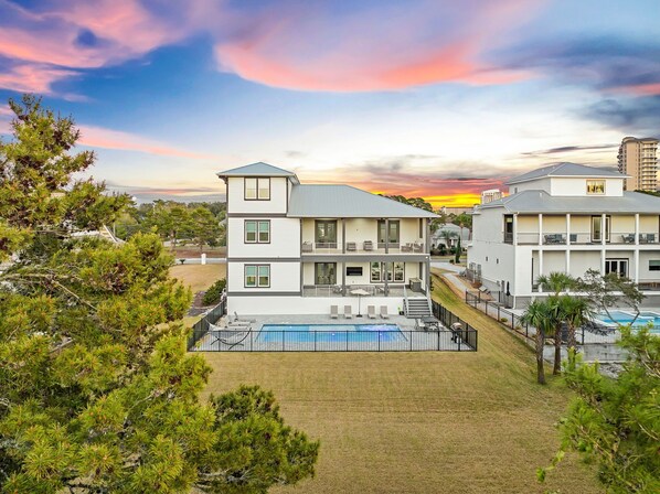 A two-story building with a swimming pool and outdoor seating, surrounded by greenery and under a sunset sky.