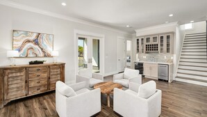 A modern living room with a wooden cabinet, white armchairs, a coffee table, and a kitchen area with cabinets and a staircase. - Rising Tide | Private Pool/Spa | 6-Seater Golf Cart | Private Beach Access (Miramar Beach)